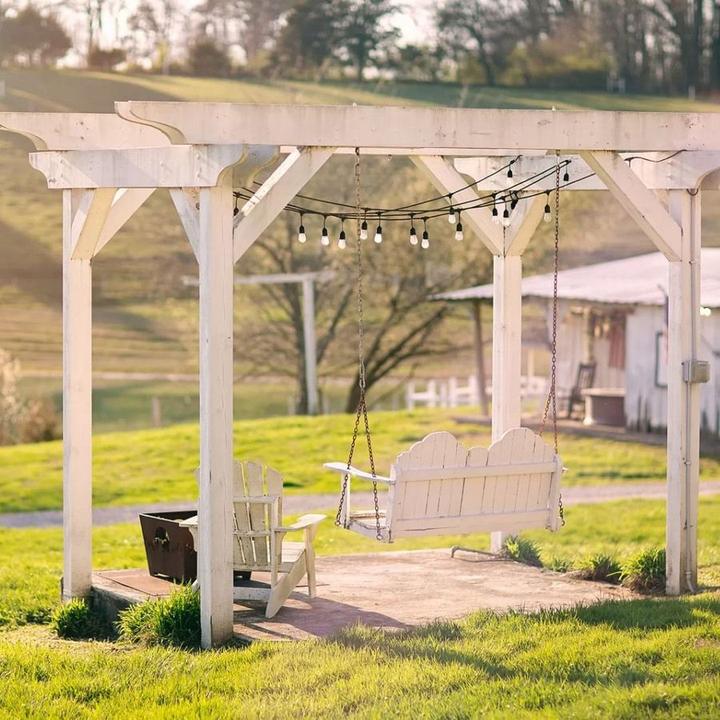 A white wooden pergola with string lights frames a porch swing overlooking a grassy field and tree line.